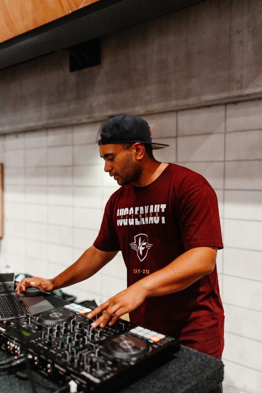 DJ Malix mixing on a DJ controller in a maroon Juggernaut Fitness t-shirt and black cap