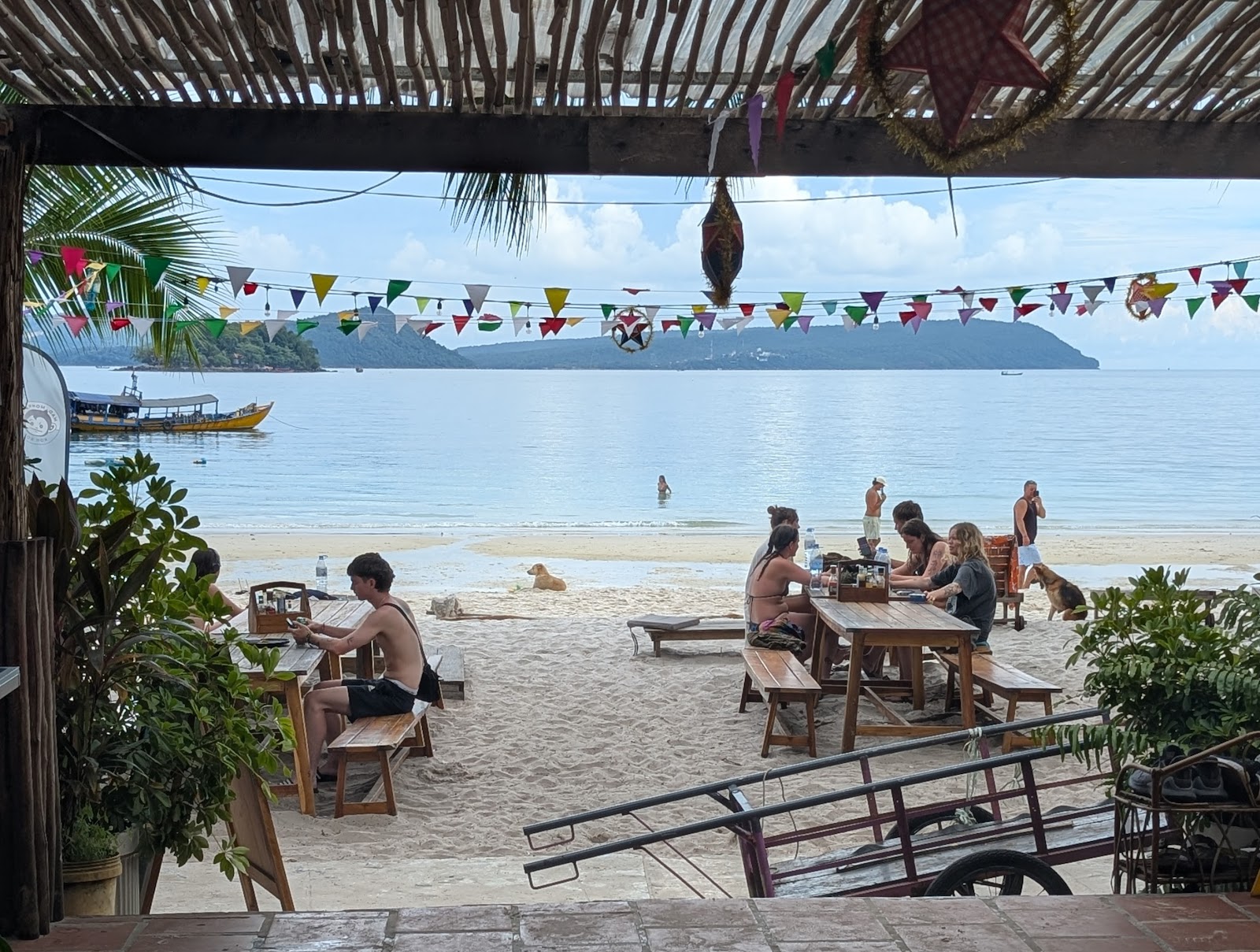 A view out from a tiled platform, over some picnic tables with
                 resting travelers, to the beach where a dog is lounging and
                 some more travelers are swimming. A few cables hang from a
                 beam overhead, with some flags and string lights over the
                 picnic tables