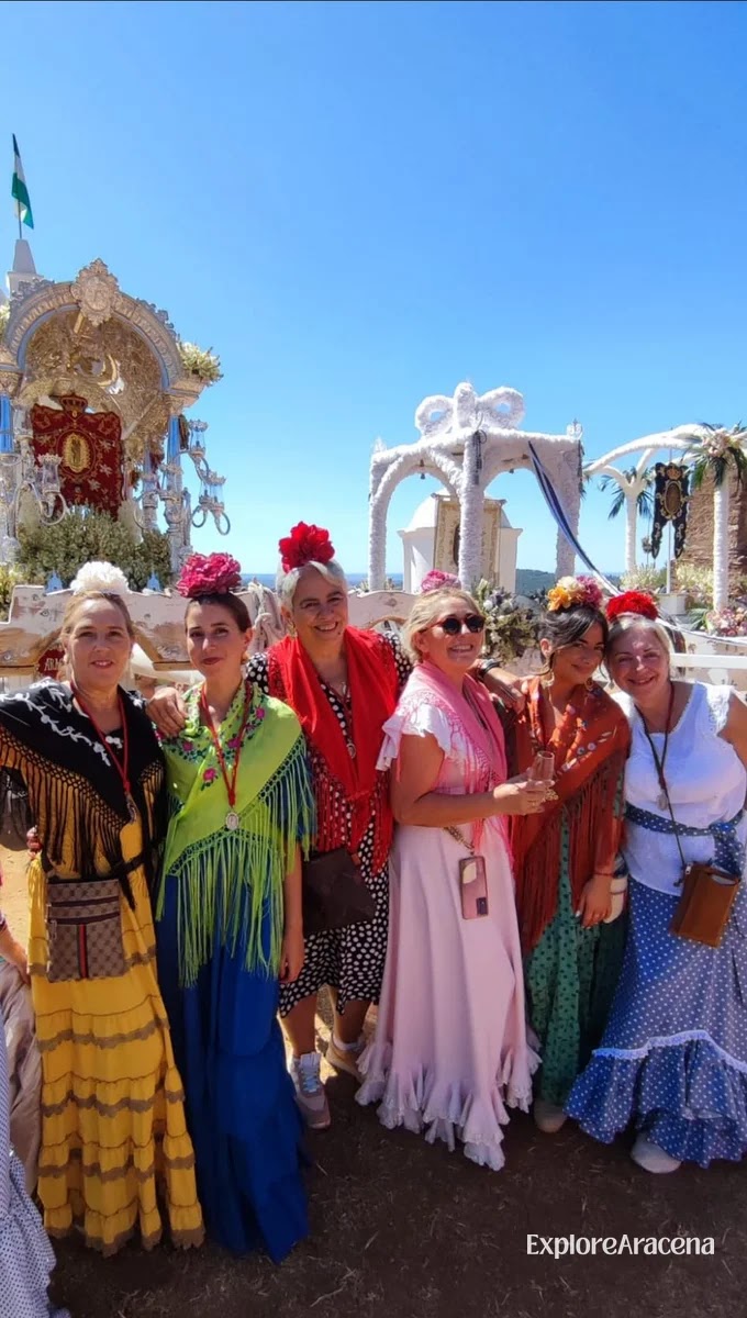 Women in flamenca dresses standing beneath floral arches at the Peña de Arias Montano on a clear day