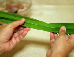 Khi thịt gà đã ngấm gia vị, lấy lá dứa, lật úp mặt lá bóng xuống dưới, mặt không bóng ngửa lên trên. Gấp chéo hai đầu lá lên trên, để 1 đầu dài hơn.