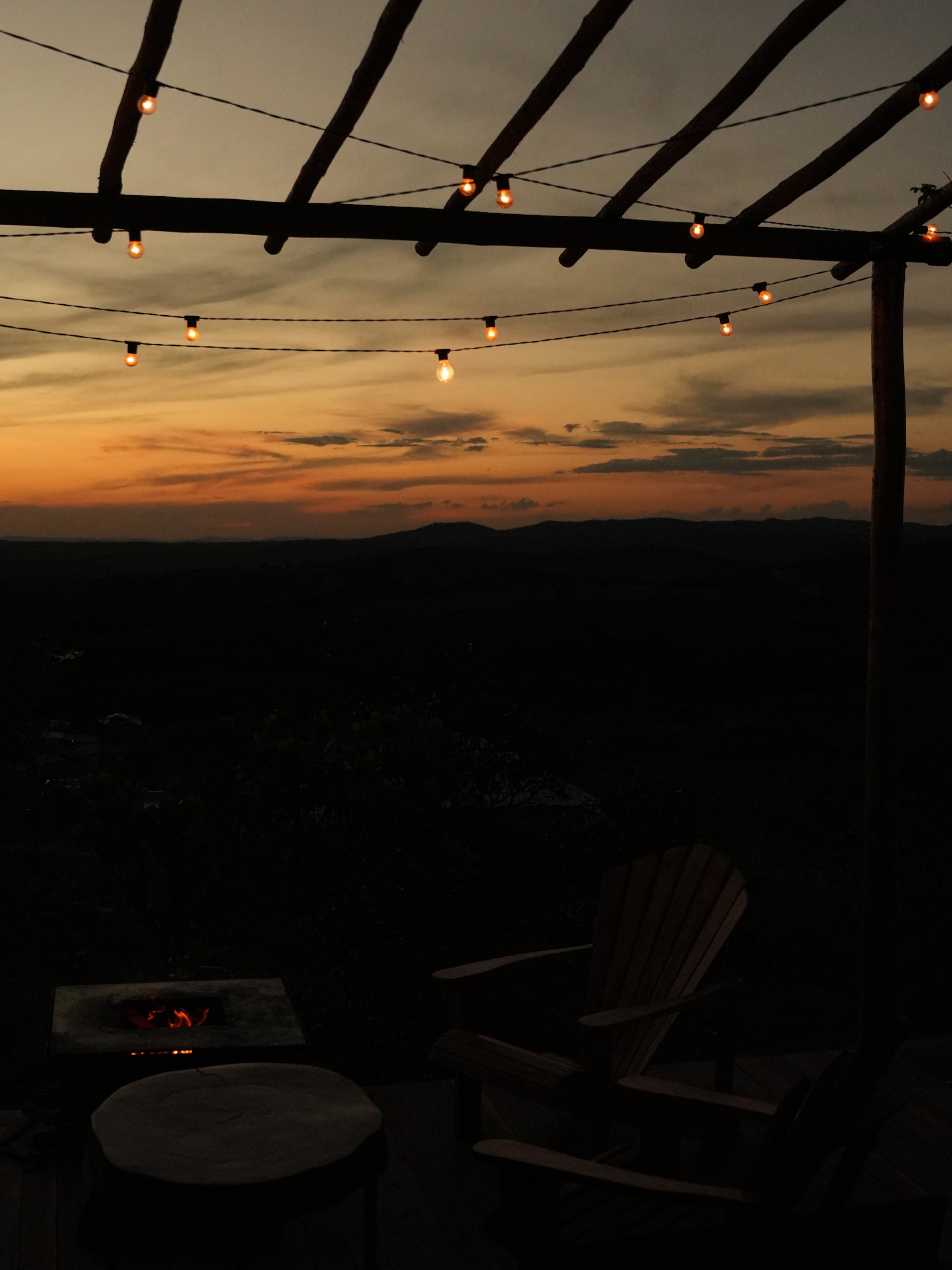 Foto real da pérgola do deck de um chalé Mei do Mato ao entardecer, com varal de lâmpadas acesas e o horizonte alaranjado da Serra do Cipó
