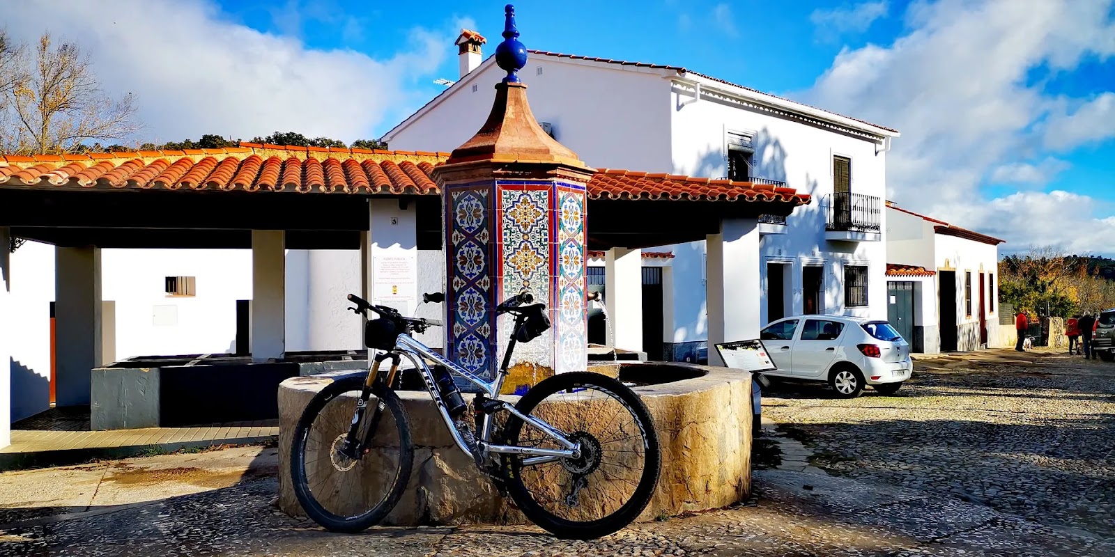 A mountain bike leaning against a stone fountain with a colourful tiled column, beside a covered public wash house in a white village of the Sierra de Aracena.