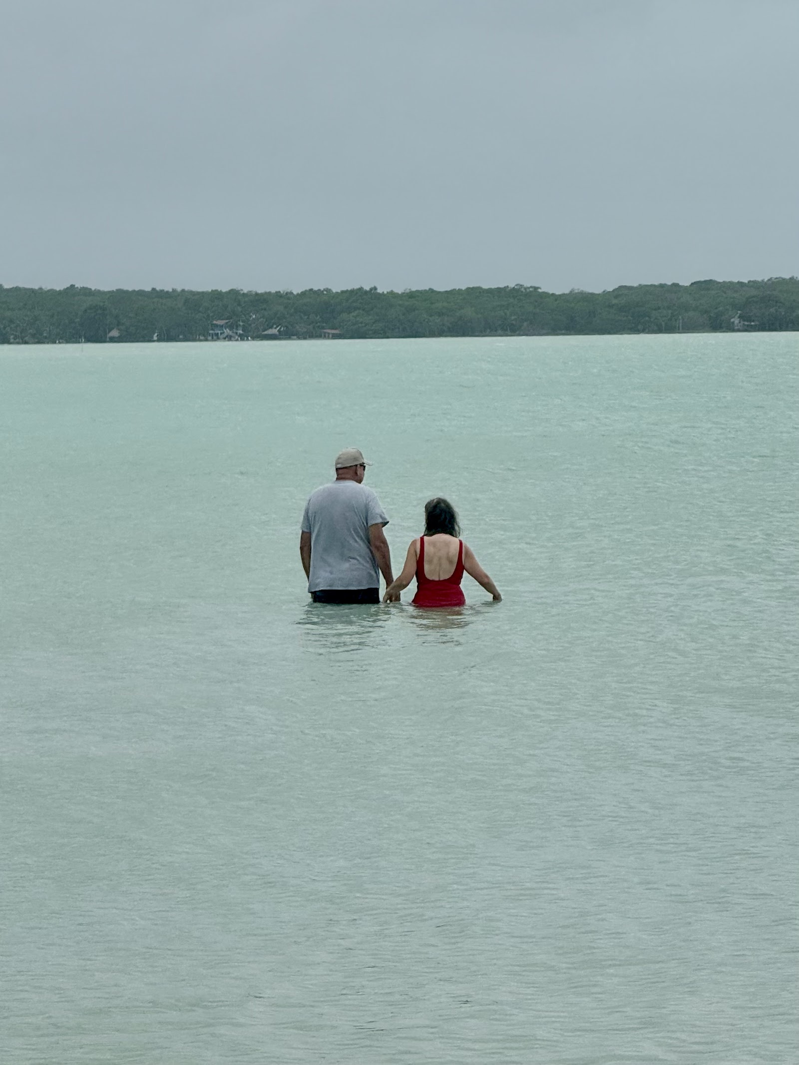A man and a woman wading out into still water in Belize, seen from the shore