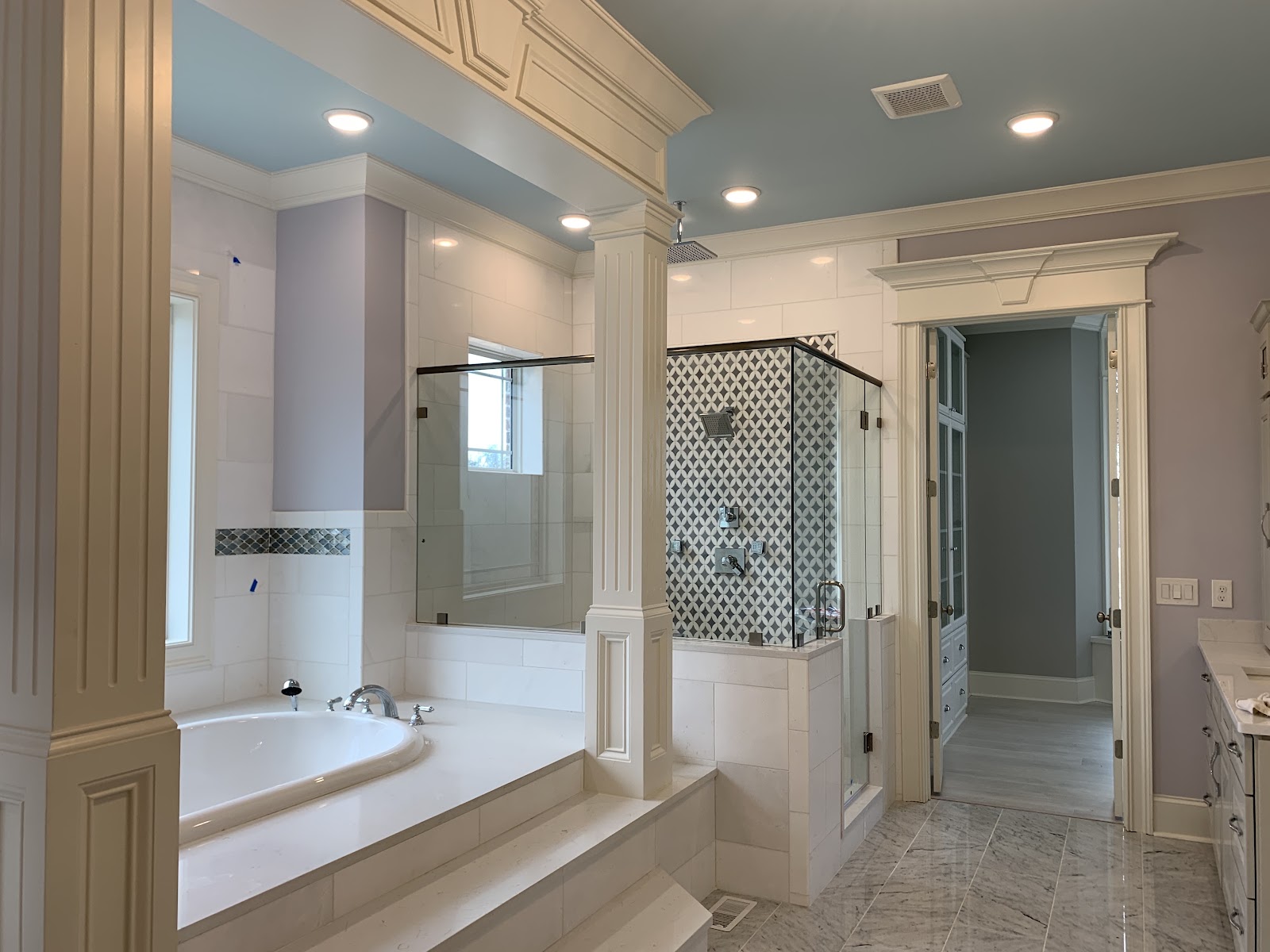 Bathroom interior featuring a white built-in bathtub, a glass-enclosed shower with geometric patterned tile, white pillars, and a light blue ceiling with recessed lighting.