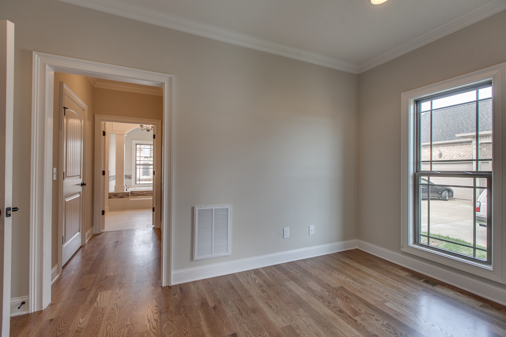 An empty room with hardwood floors, a window, and an open doorway leading to a hallway and bathroom.