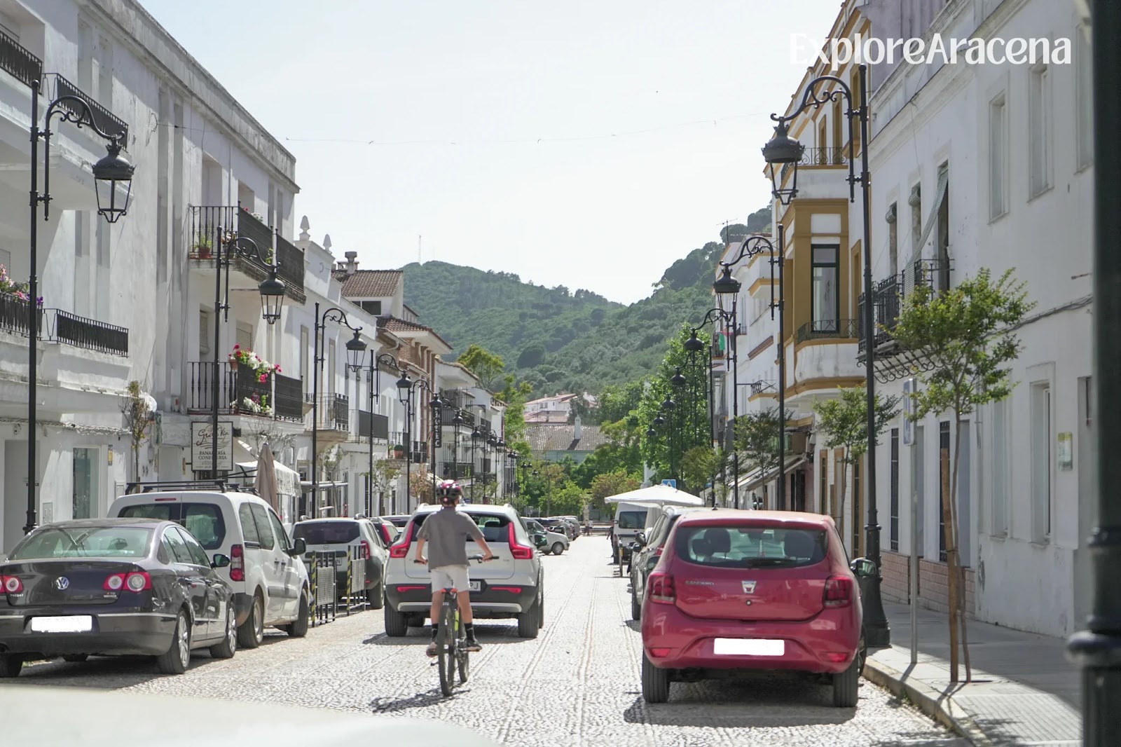 A cyclist riding through a street in Aracena, with white buildings and lush green vegetation in the background.