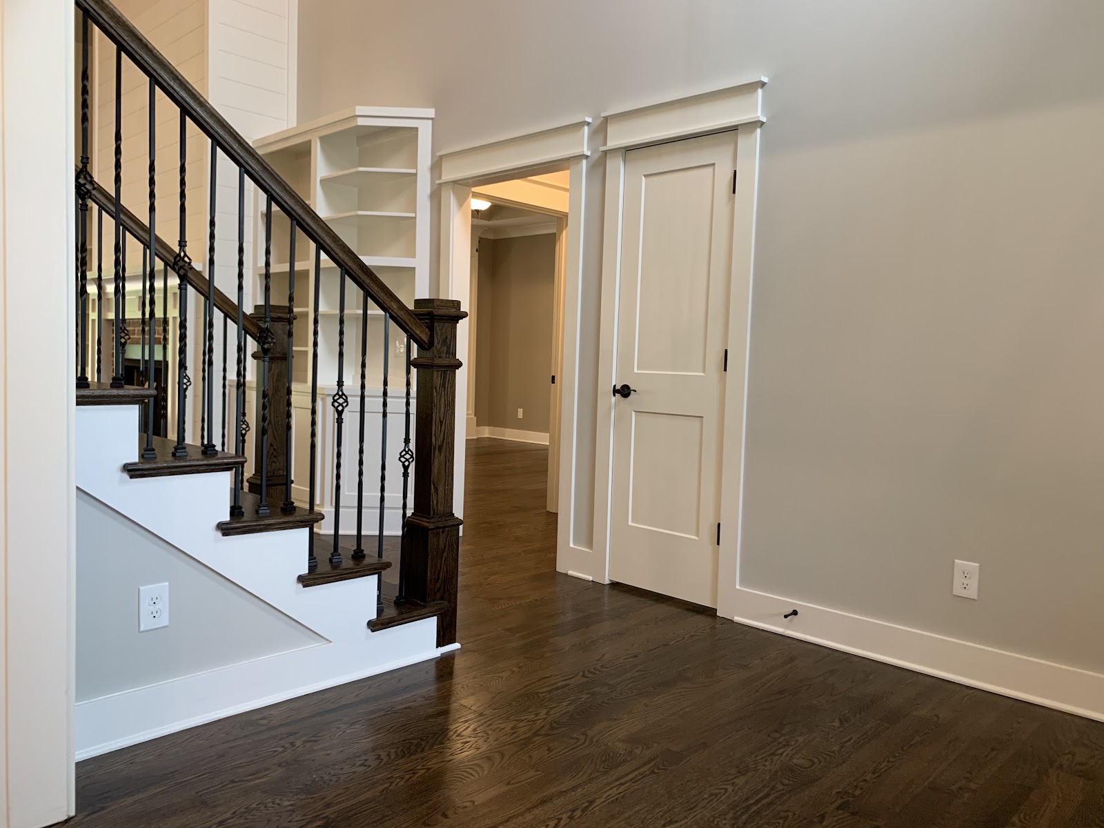 Interior view of a home featuring a dark wood staircase with black metal spindles, white built-in shelving, and a white door. The space has neutral gray walls and dark wood flooring.
