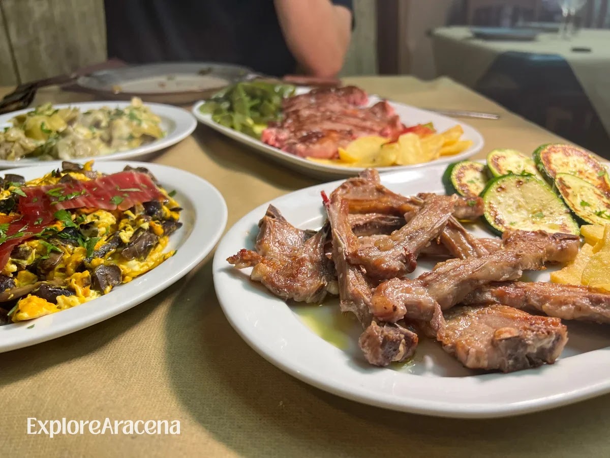 Table spread at Restaurante José Vicente showing grilled lamb ribs with courgette and potatoes, scrambled eggs with mushrooms and jamón, and a mixed grilled meat platter