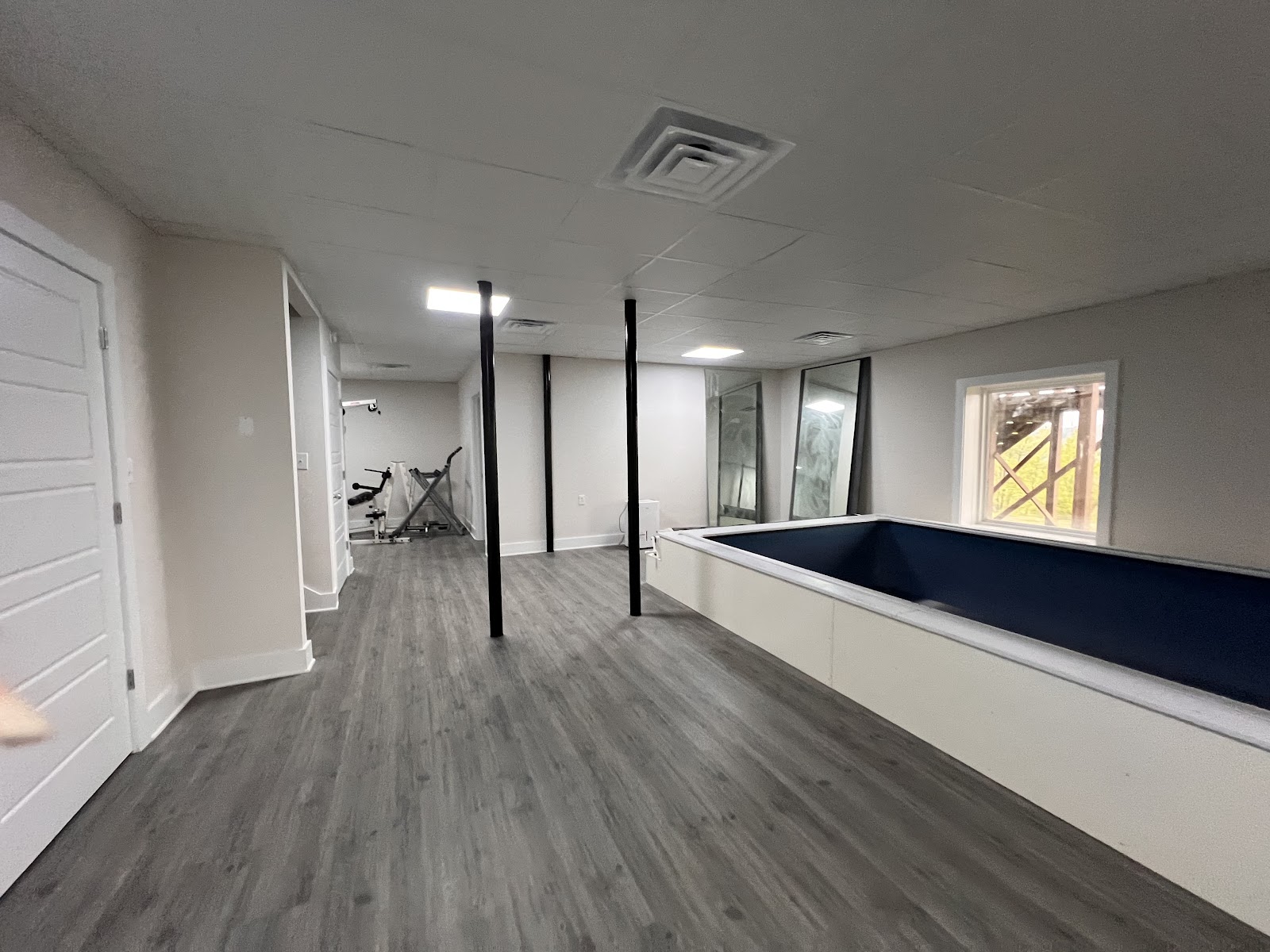 Indoor room with grey wood-look flooring, black support columns, and a raised rectangular tank with a dark blue interior. Exercise equipment is visible in the background.