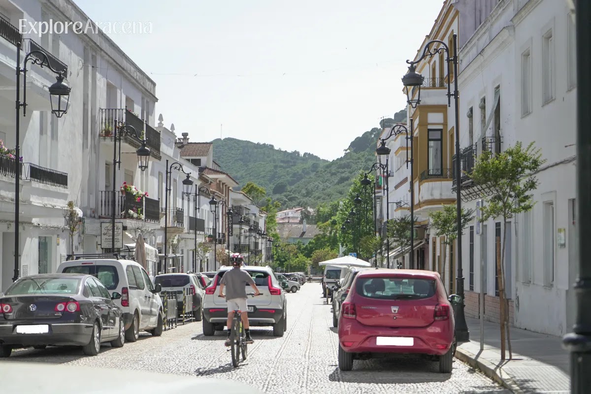 Cyclist riding along a road in Aracena with white buildings, a passing car, and green hills in the background.