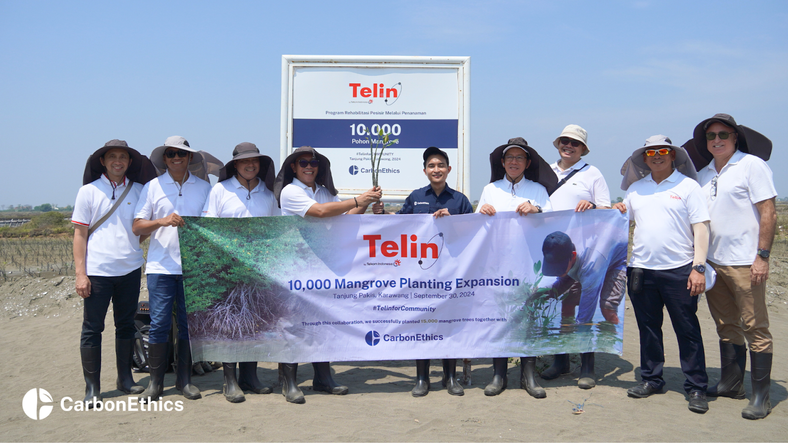 Group photo of Telin and CarbonEthics representatives standing together holding a banner that reads '10,000 Mangrove Planting Expansion' at a mangrove restoration site in Tanjungpakis, Karawang, West Java, with a project signboard in the background.