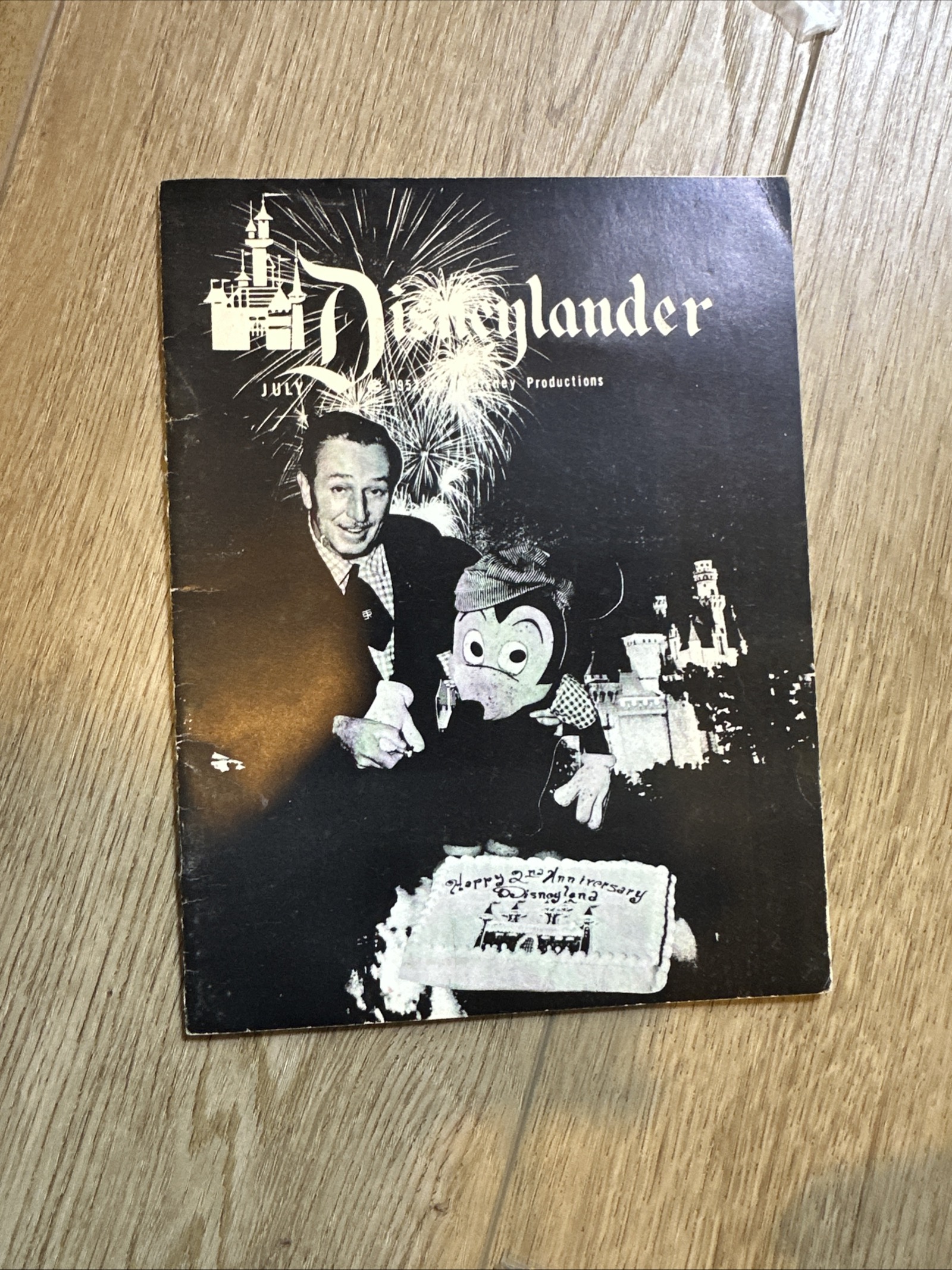 Black and white cover of The Disneylander July 1957 employee newsletter showing Walt Disney and costumed Mickey Mouse behind an anniversary cake with fireworks over Sleeping Beauty Castle