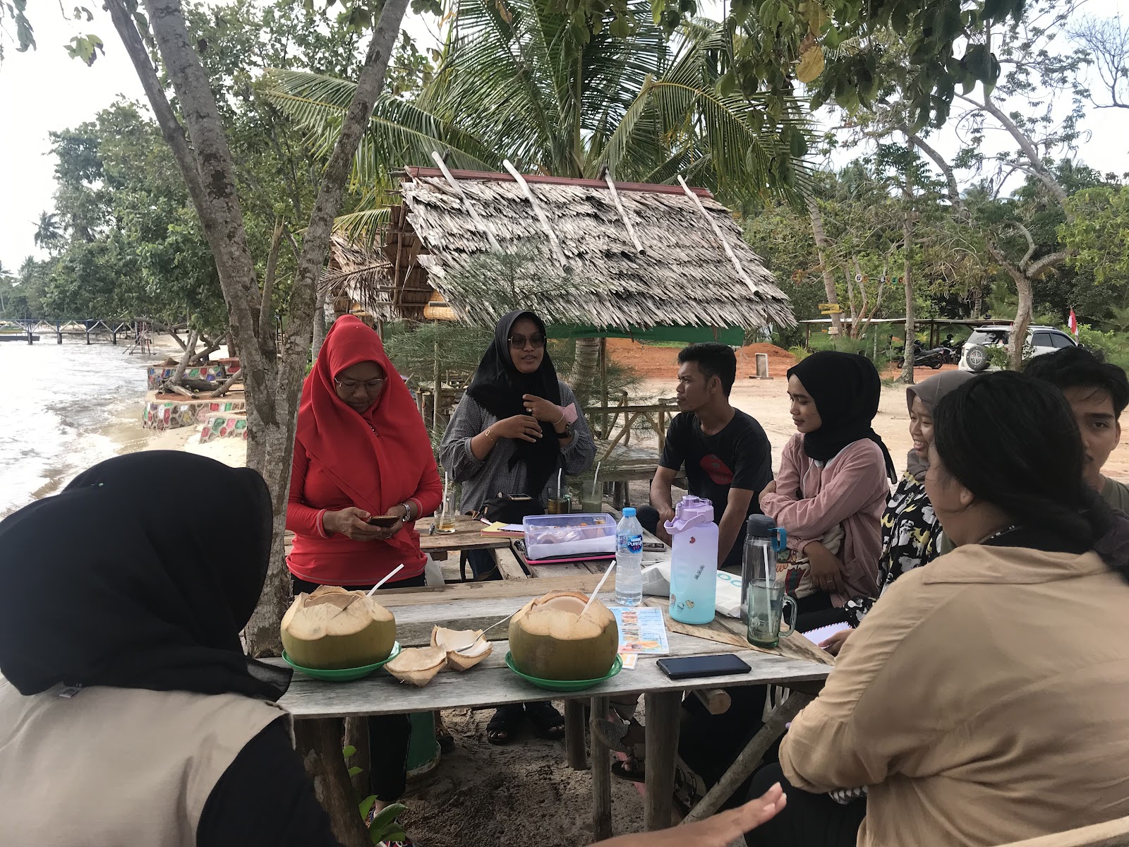Villagers and teenagers bonding under a tree sipping coconut water on the beach. Dompak island, blue carbon education, Indonesia coastal communities. CarbonEthics Indonesia