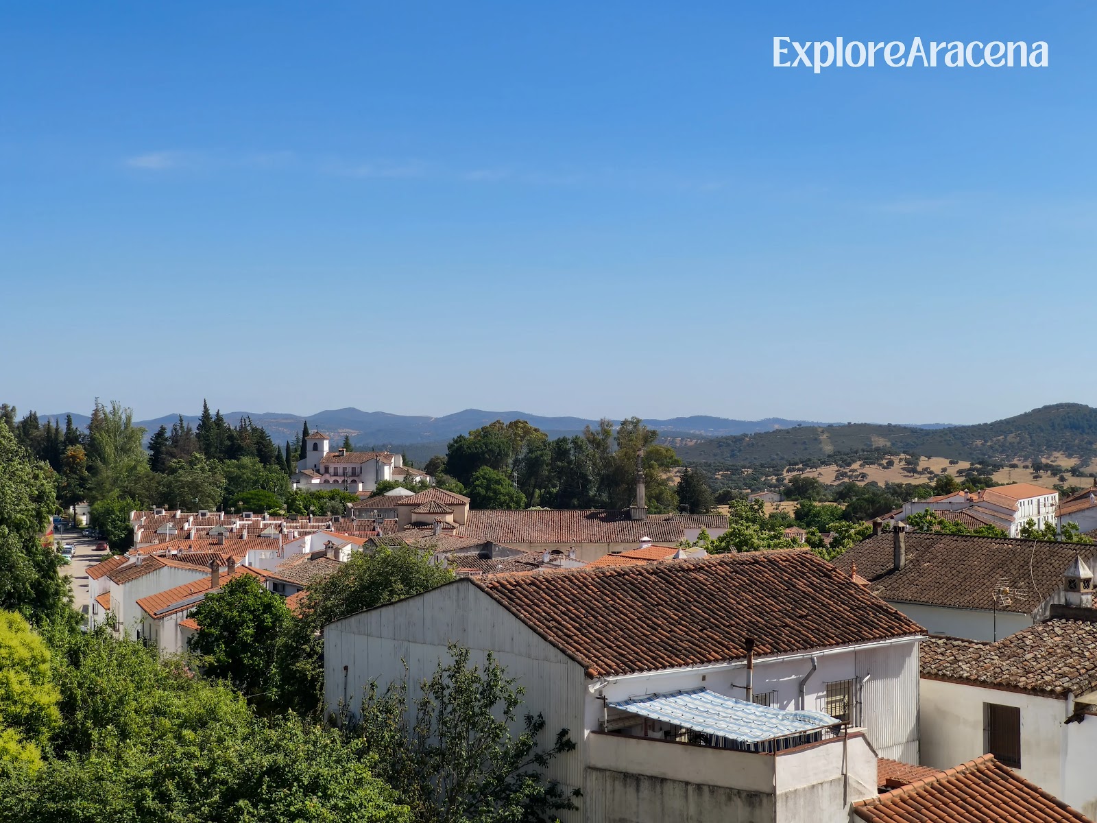 White houses in Aracena surrounded by trees, with wooded hills beneath a blue sky.