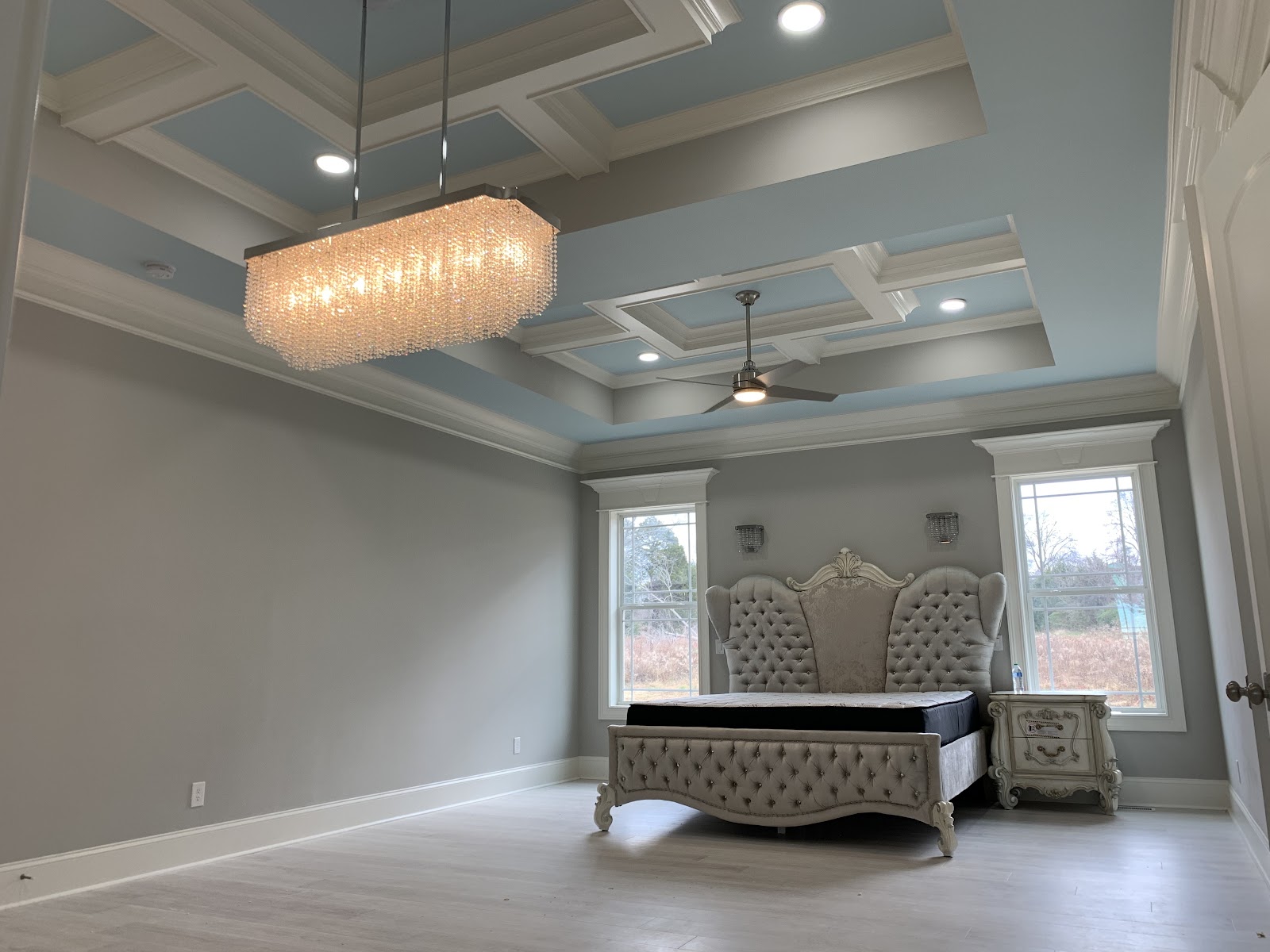 Bedroom with a tufted headboard, light wood flooring, and a coffered ceiling with blue panels. A large rectangular crystal chandelier hangs above the bed.