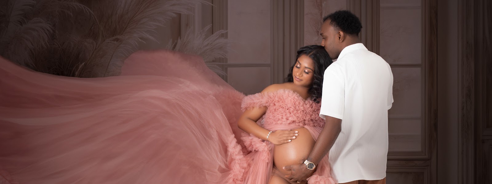 Studio maternity portrait of a couple: expectant mother in a flowing blush-pink tulle gown; partner cradles her belly against an elegant backdrop