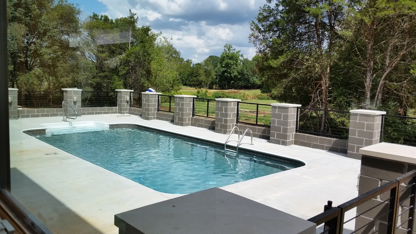 Daytime view of an outdoor swimming pool and concrete deck enclosed by a block pillar and metal railing fence at the Blocky Custom Home.
