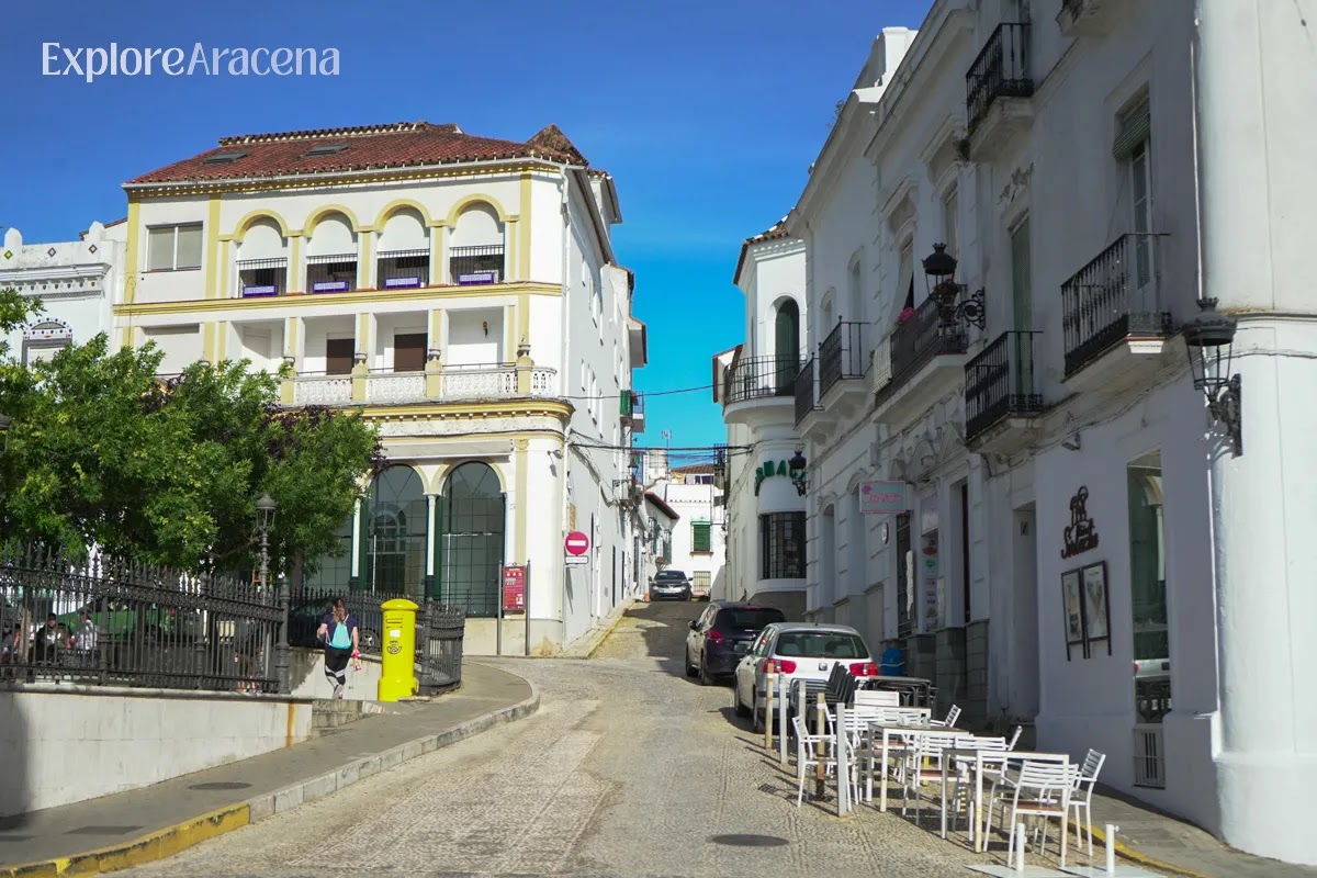 Street scene in Aracena with pedestrians, a child running along the sidewalk, white buildings, and green hills in the background.