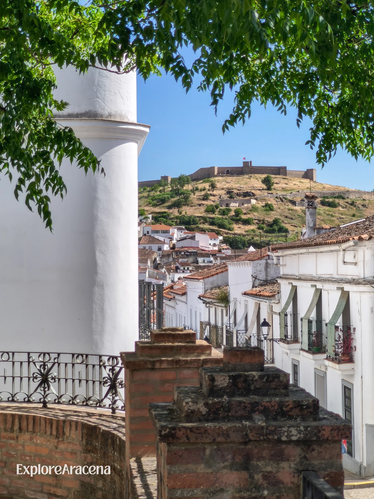 Whitewashed buildings in Aracena beneath a shady tree, with Aracena Castle visible on the hill.