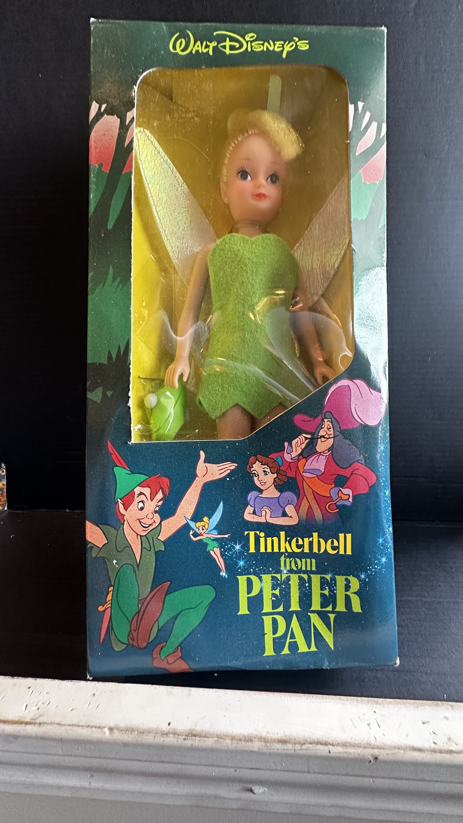 Vintage Tinker Bell fashion doll in original teal window box showing green felt dress, rooted blonde hair in bun, and translucent plastic wings, with Peter Pan character art on packaging