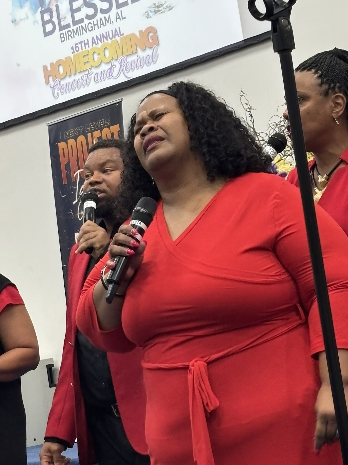 Female soloist in red dress singing at the Homecoming Concert with BLESSED banner in background