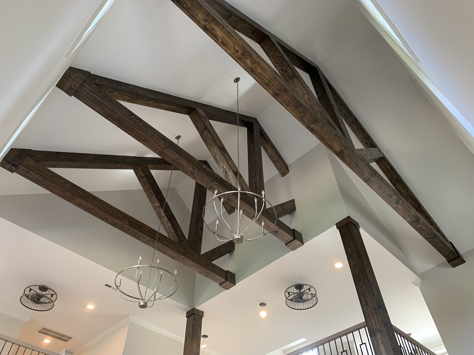 Upward view of a vaulted white ceiling featuring large, dark-stained wood trusses and two circular metal chandeliers.