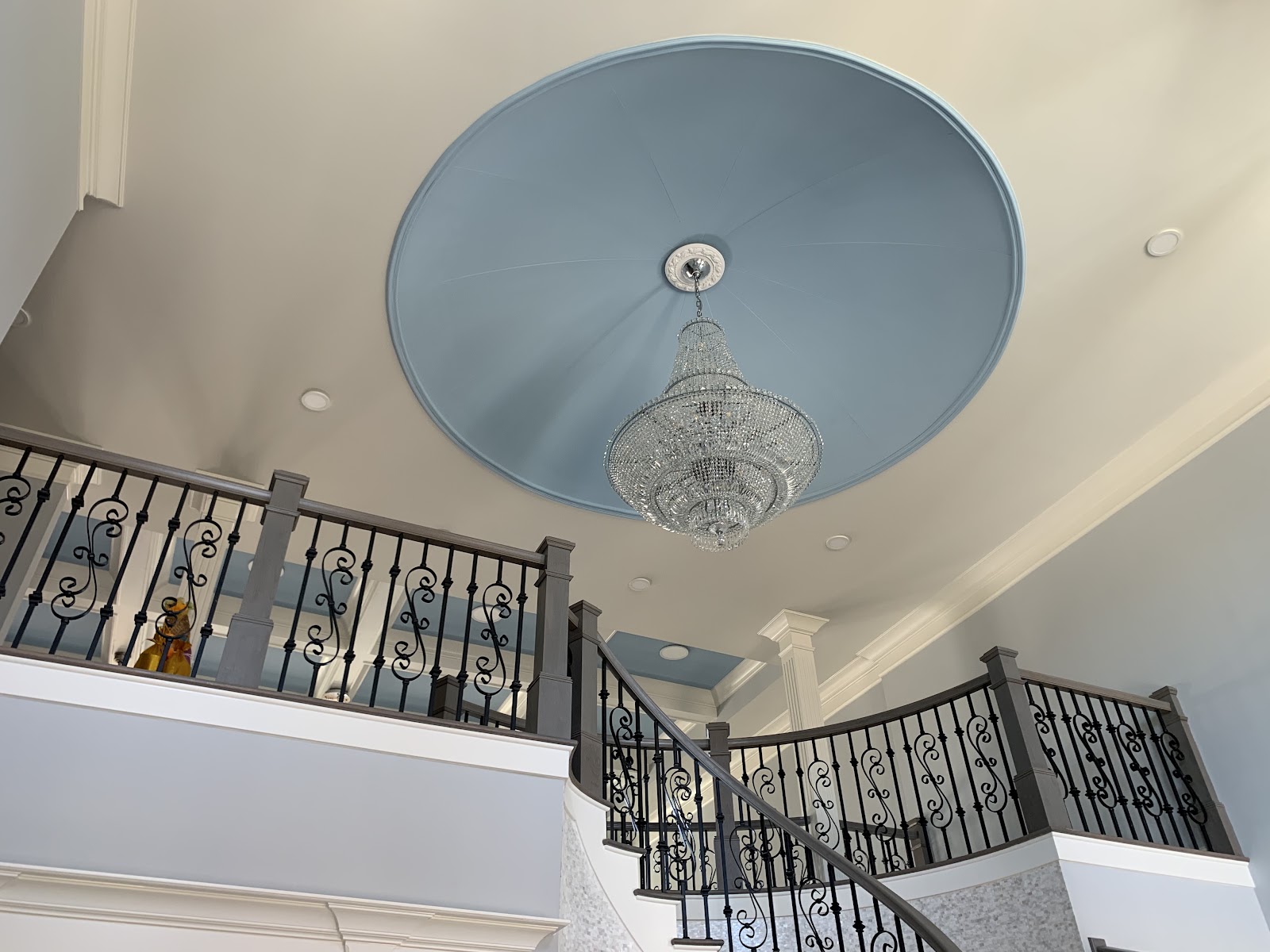 Low-angle view of a large crystal chandelier hanging from a circular blue ceiling tray. A curved staircase with dark wood handrails and decorative wrought iron balusters is visible below.