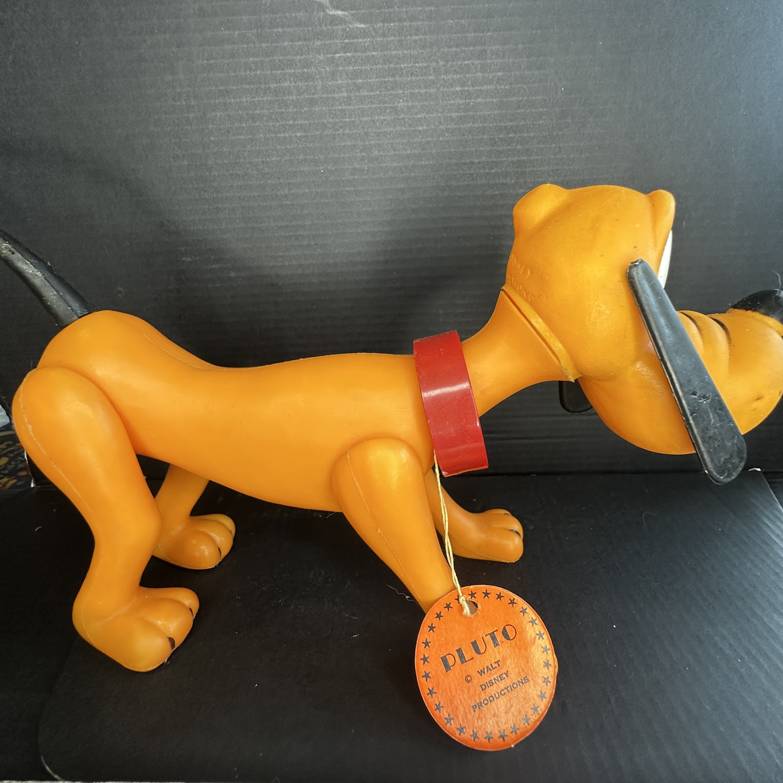 Vintage yellow-orange soft vinyl Pluto squeak toy by Sun Rubber Co., standing on four legs with red collar, long black ears, and original circular orange hang tag attached to front leg