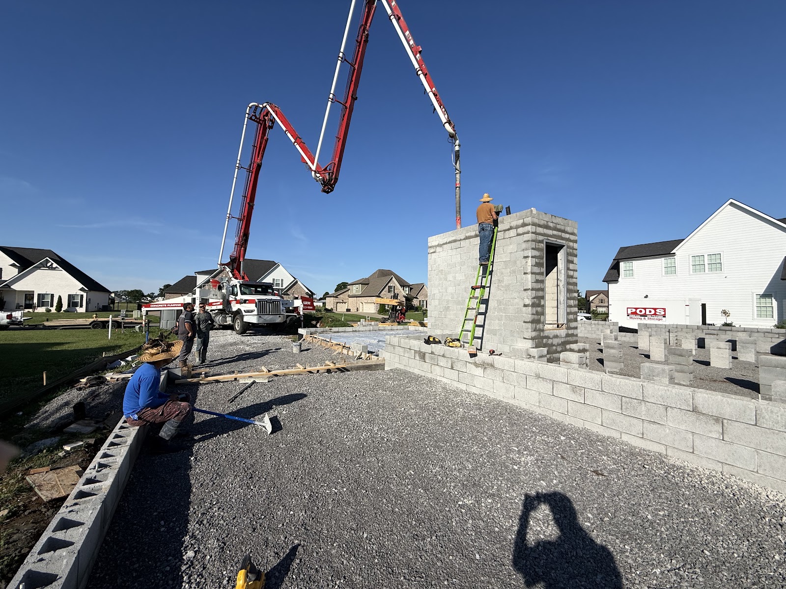 A concrete pump truck boom extends over a concrete block storm shelter under construction at a residential site.