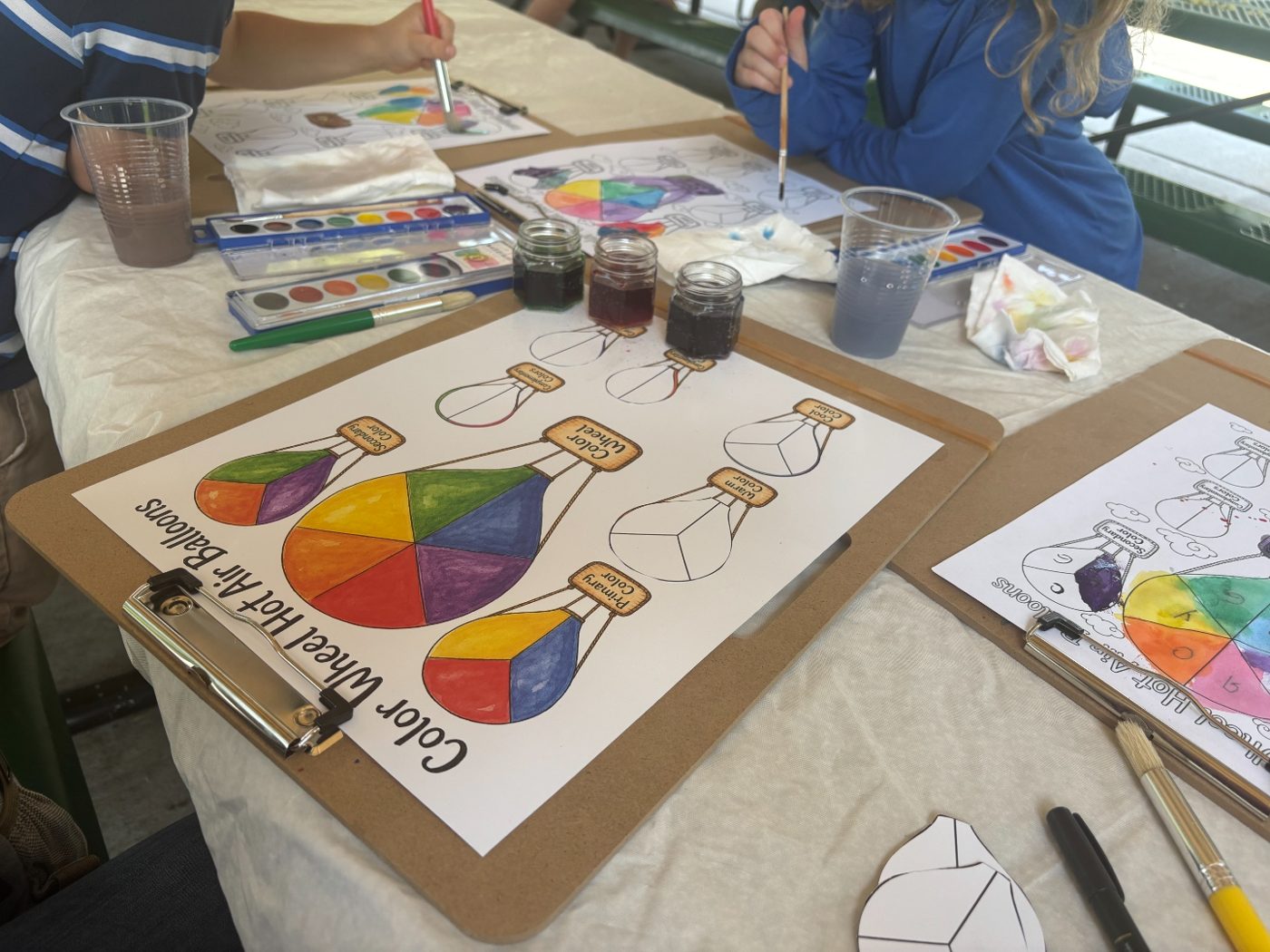 Children at a picnic table painting color-wheel hot air balloon worksheets with watercolors