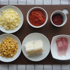 Đun nóng 2 muỗng canh dầu ăn, cho hành tây băm và tỏi băm vào đảo đều 1 phút. Sau đó thêm vào 7 muỗng canh sốt cà chua, 4 muỗng canh siro bắp, 1 muỗng canh tương ớt Hàn Quốc, đảo đều. Khi hỗn hợp sốt sôi lên thì cho bánh gạo và thịt xông khói thái nhỏ, đun khoảng 4-5 phút cho bánh gạo chín ngấm đều sốt là được.