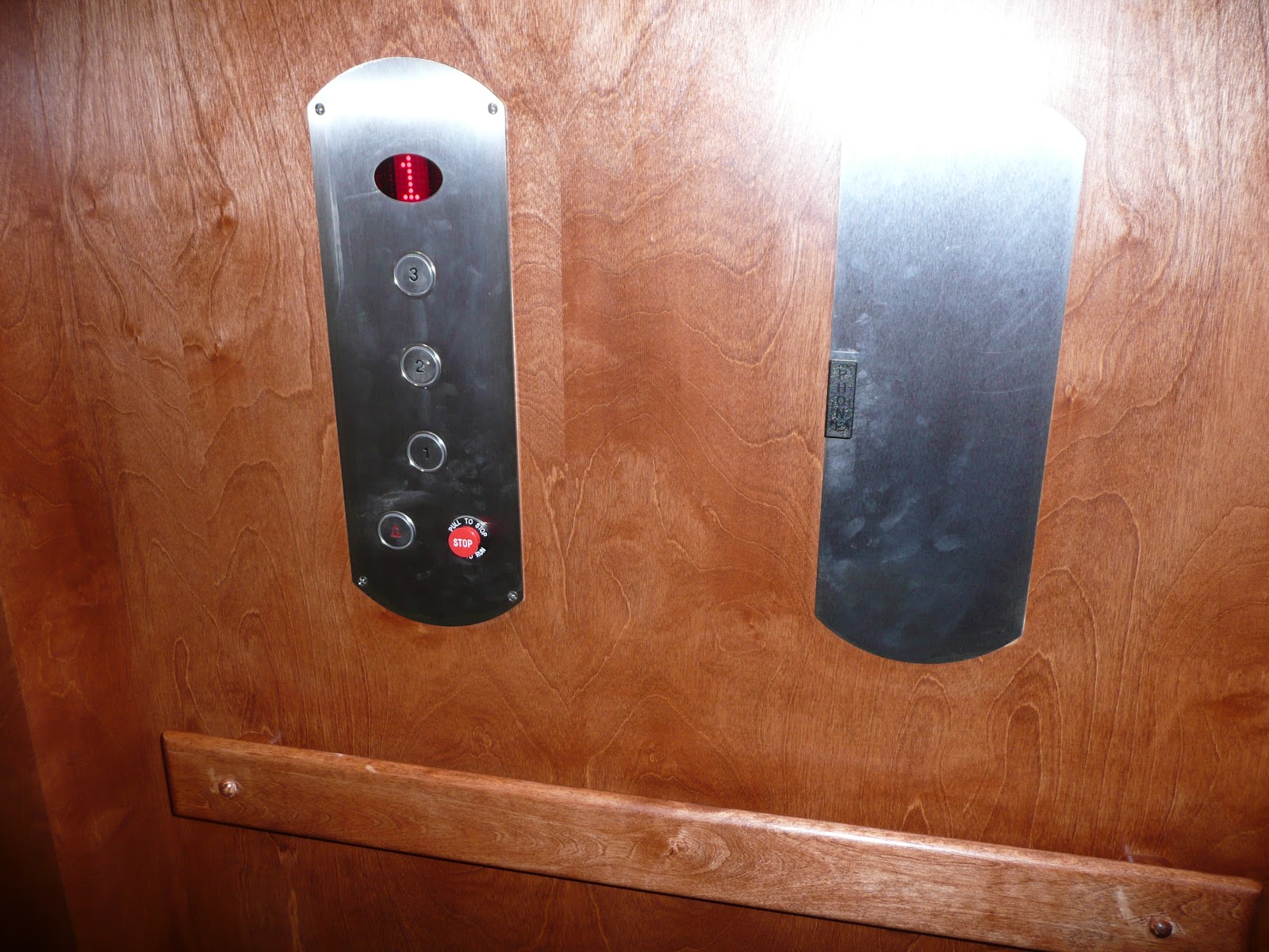Close-up of an elevator's interior control panel on a wood-grain wall, featuring floor buttons and a red stop button.