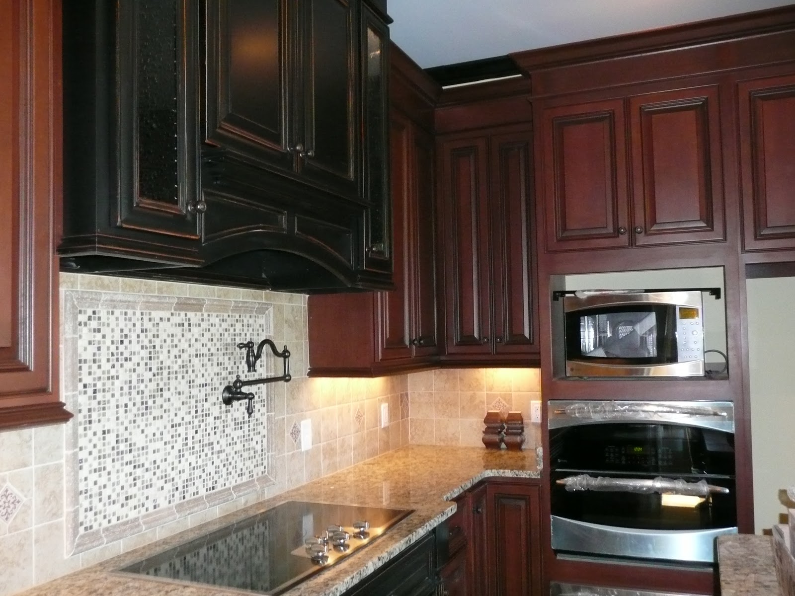 Kitchen interior with dark cabinetry, stone-patterned countertops, built-in double ovens, and a tiled backsplash with a mosaic pattern behind the stovetop.