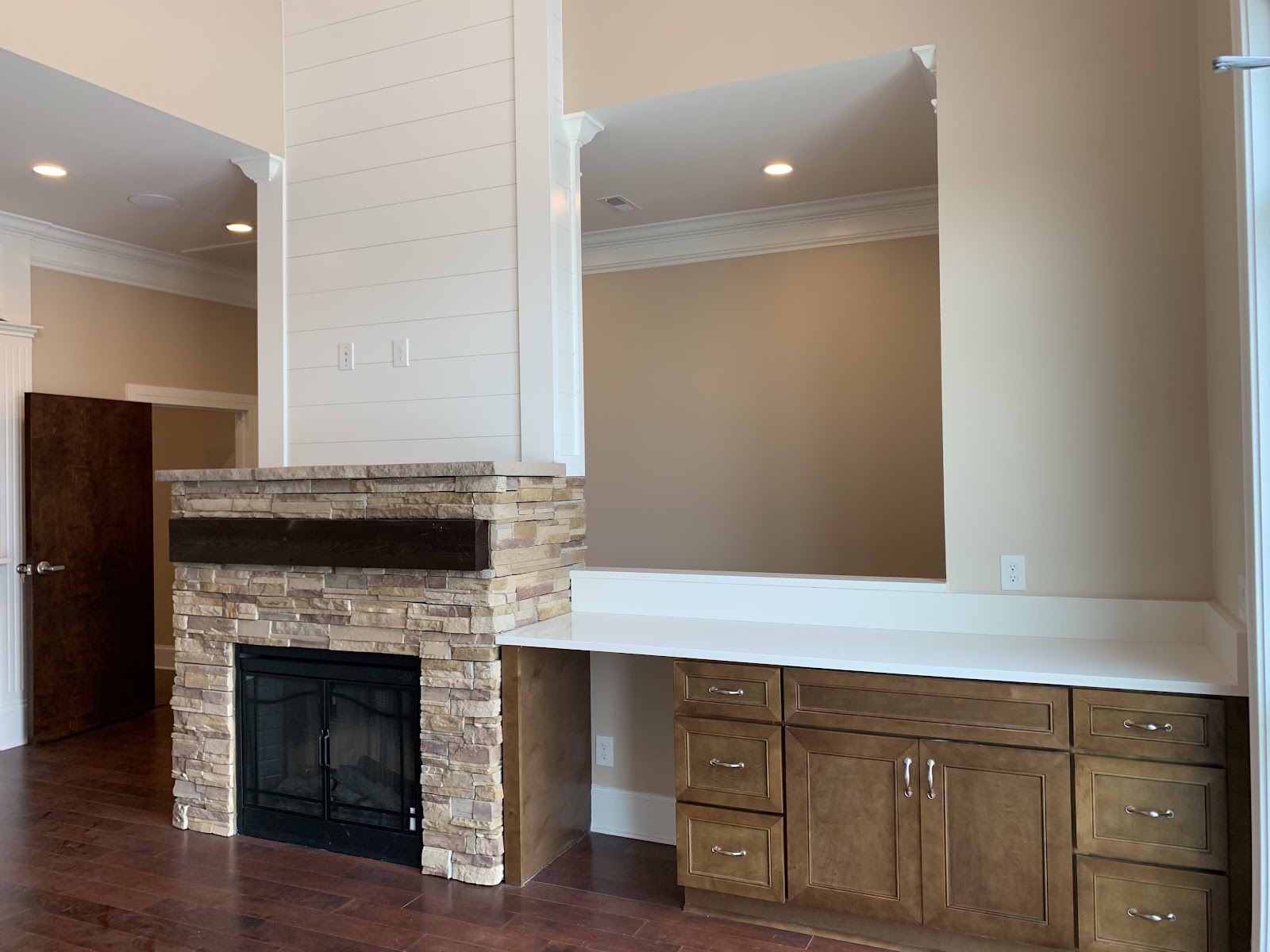 A stone-faced fireplace with a dark wood mantel and white shiplap above. Adjacent is a built-in desk with brown cabinets and a white countertop over dark wood flooring.