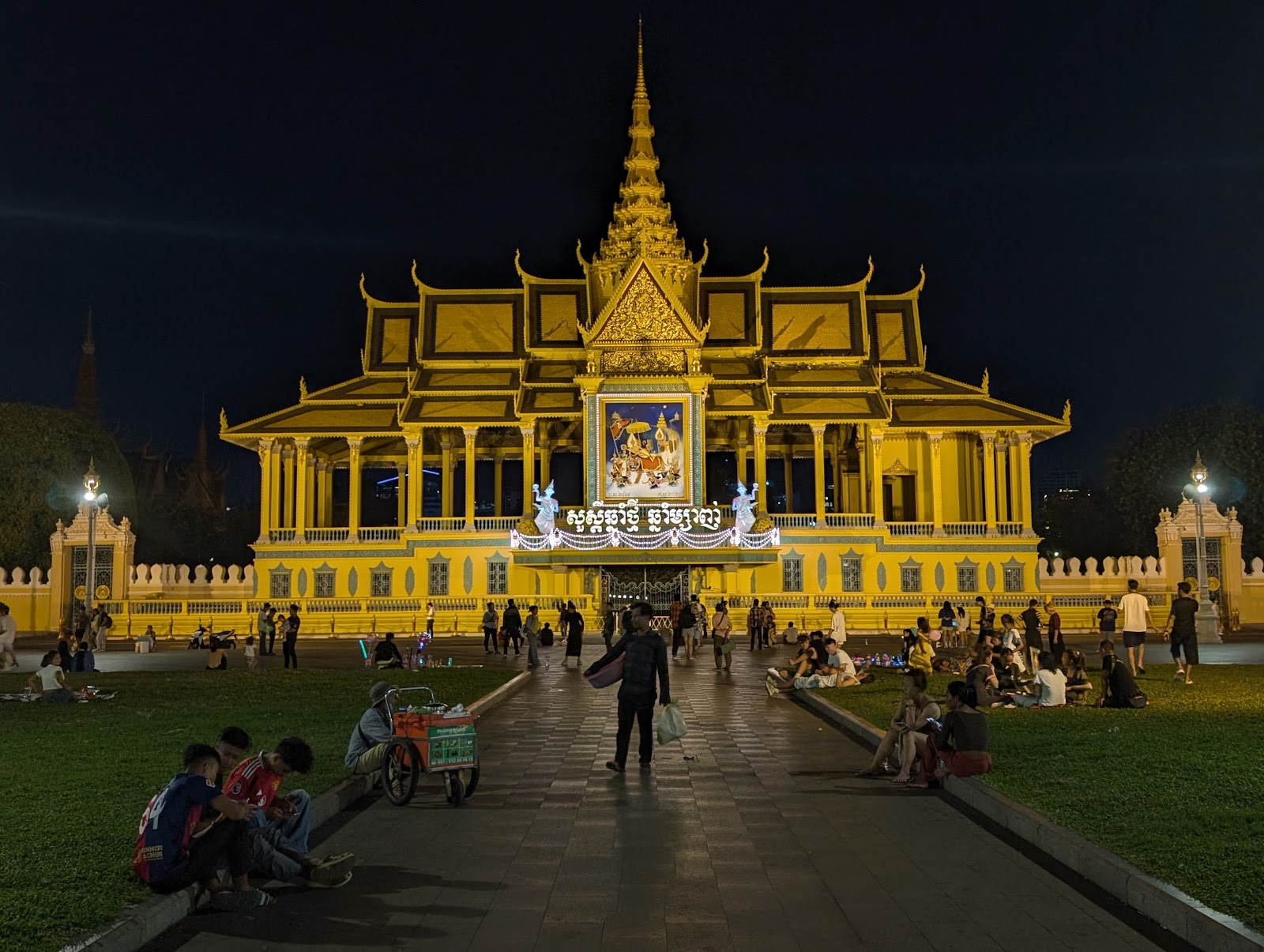 The Cambodian Royal Palace lit up in yellow light at night in
                 front of a park with a wide paverstone path filled with people
                 walking and sitting on the edge, enjoying each others' company