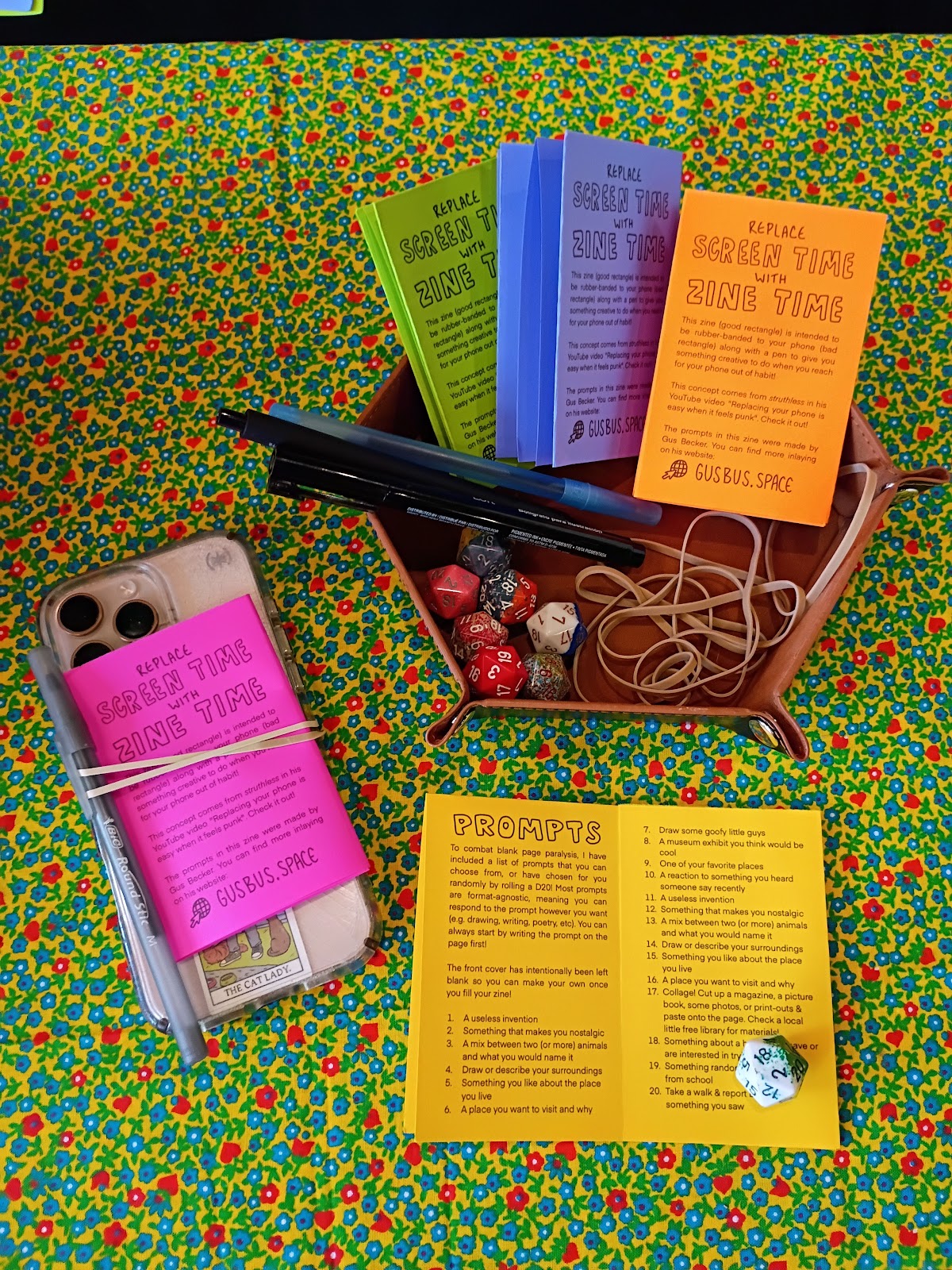 Photo showing a minizine rubber-banded to a cellphone with
                 a pen beside a bowl of more zines, pens, and 20-sided dice.
                 One of the zines lies open on the table, showing the 20
                 prompts inside
