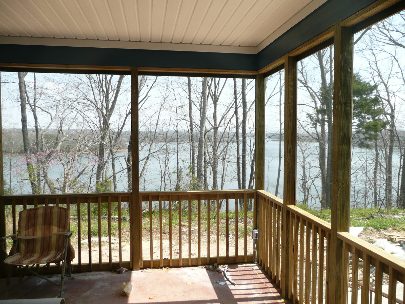 View from inside a screened-in porch with wooden railings, looking out at bare trees and a body of water. A single striped chair sits on the porch.