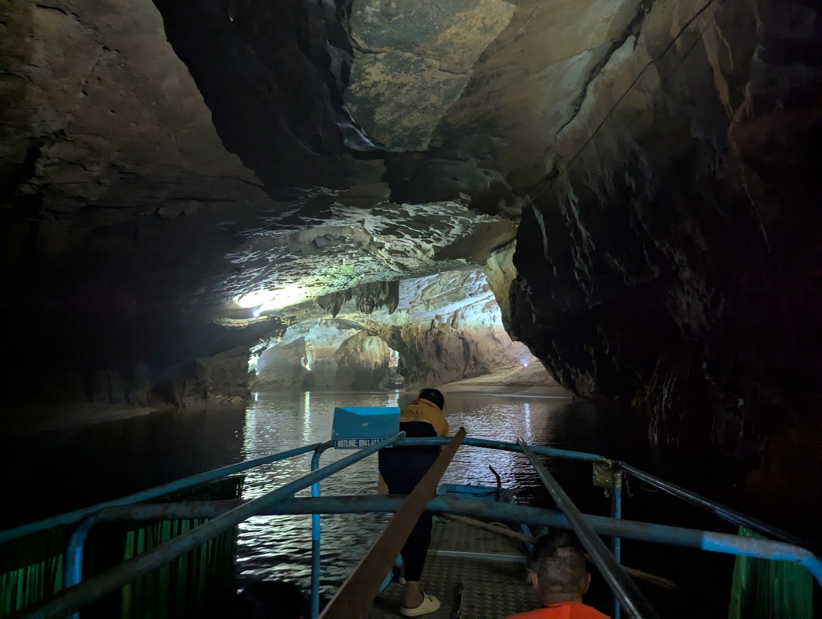 A dark photo of a person standing at the front of a boat, leaning forward
                 mid-paddle. To all side of the boat, cave features are illuminated and the
                 reflections bounce of the surface of the still water.