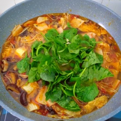 Khi nước lẩu sôi thì cho lần lượt nấm - cải thảo - giá - chả cá - bánh gạo vào. Nêm nếm lại cho vừa ăn. Dọn ra bàn ăn, cho miến + rau mồng tơi vào và thưởng thức.