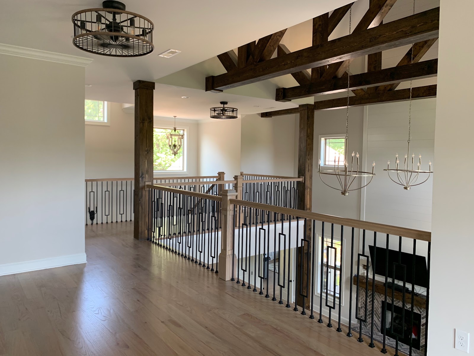 View from a second-floor landing with wood flooring and metal railings, looking toward a high ceiling with exposed dark wood trusses and chandeliers.