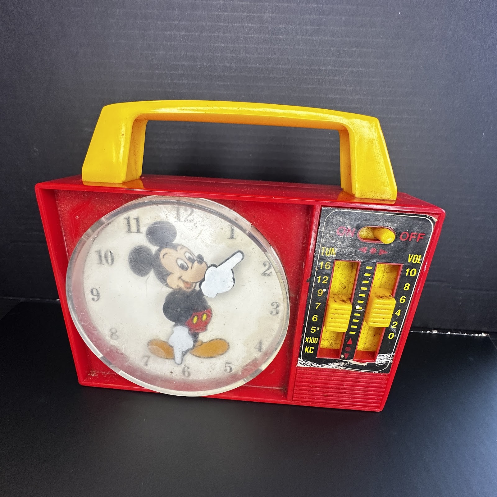 1970s Mickey Mouse AM transistor radio with red plastic body, yellow handle and sliders, and a clock-face tuning dial featuring Mickey's gloved hands as frequency pointers