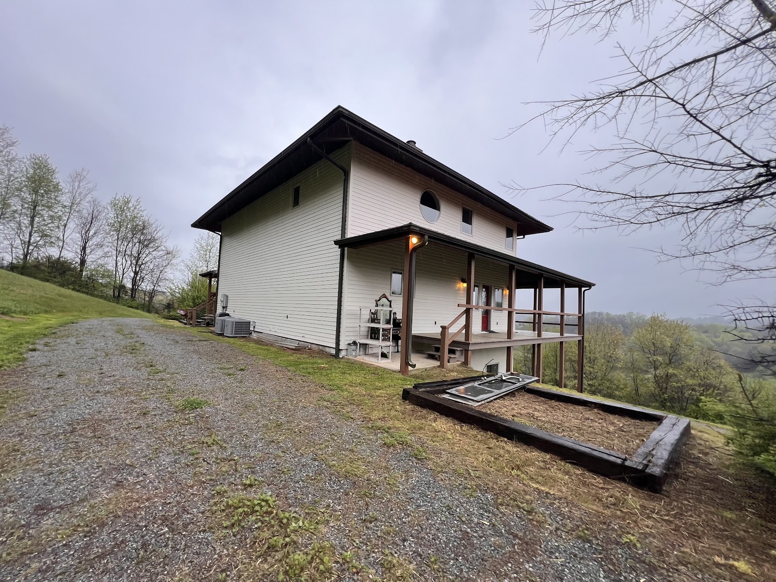 Exterior of a two-story house with light siding and a large wooden deck, situated on a hillside with a gravel driveway.