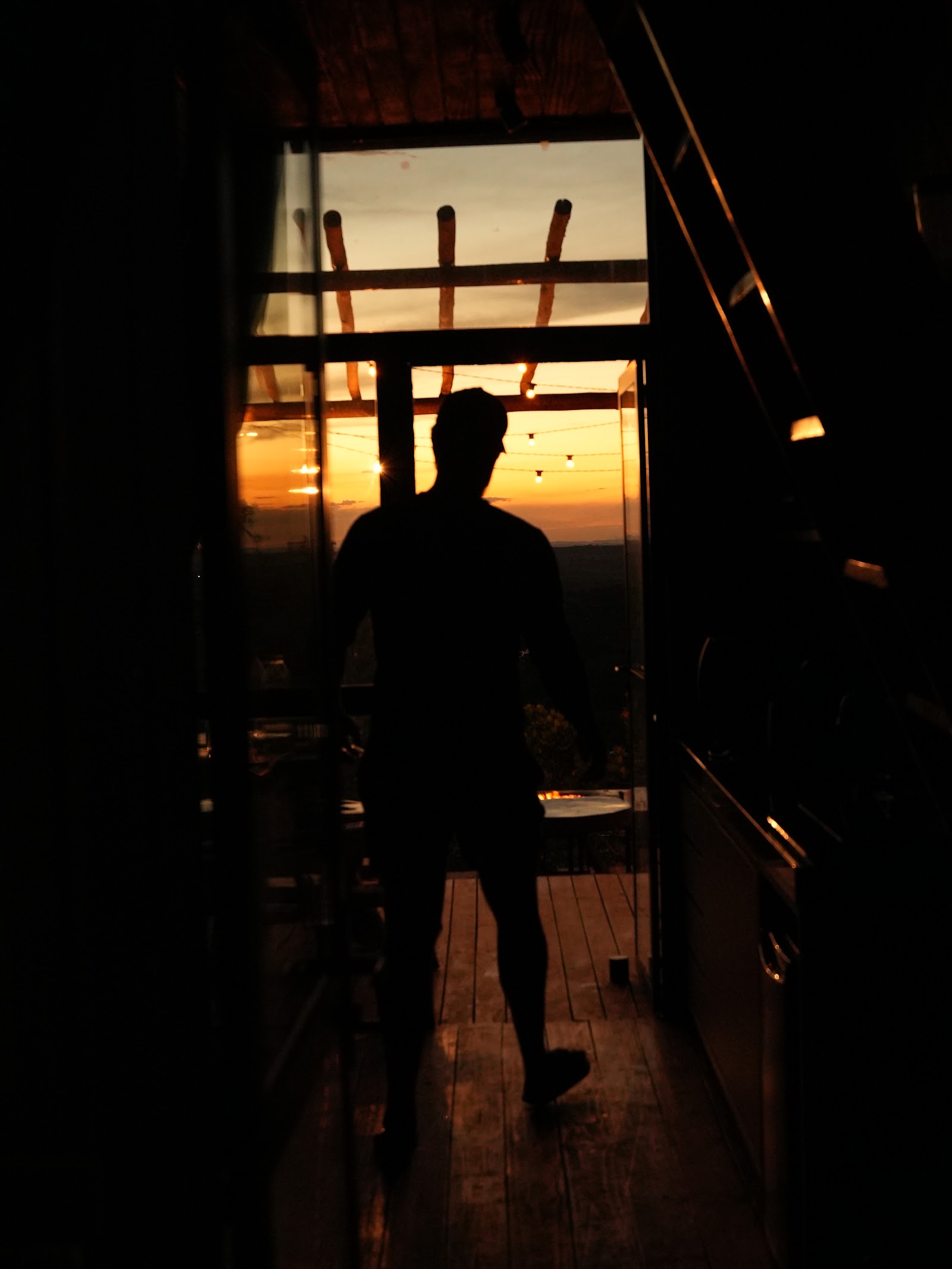 Foto real de uma pessoa atravessando a sala de um chalé A-frame da Mei do Mato ao entardecer, com o pôr do sol emoldurado pela fachada de vidro