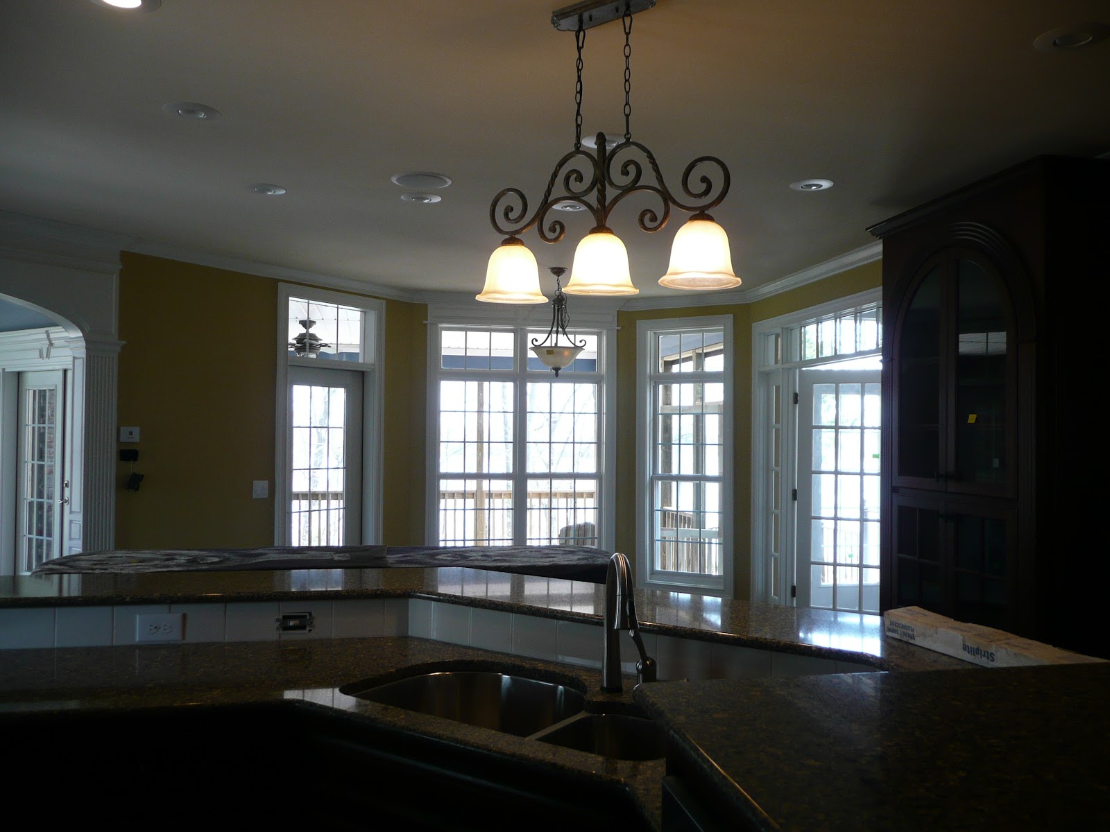 Interior view from a kitchen counter showing a stainless steel sink, dark countertops, a decorative three-bulb chandelier, and large windows.