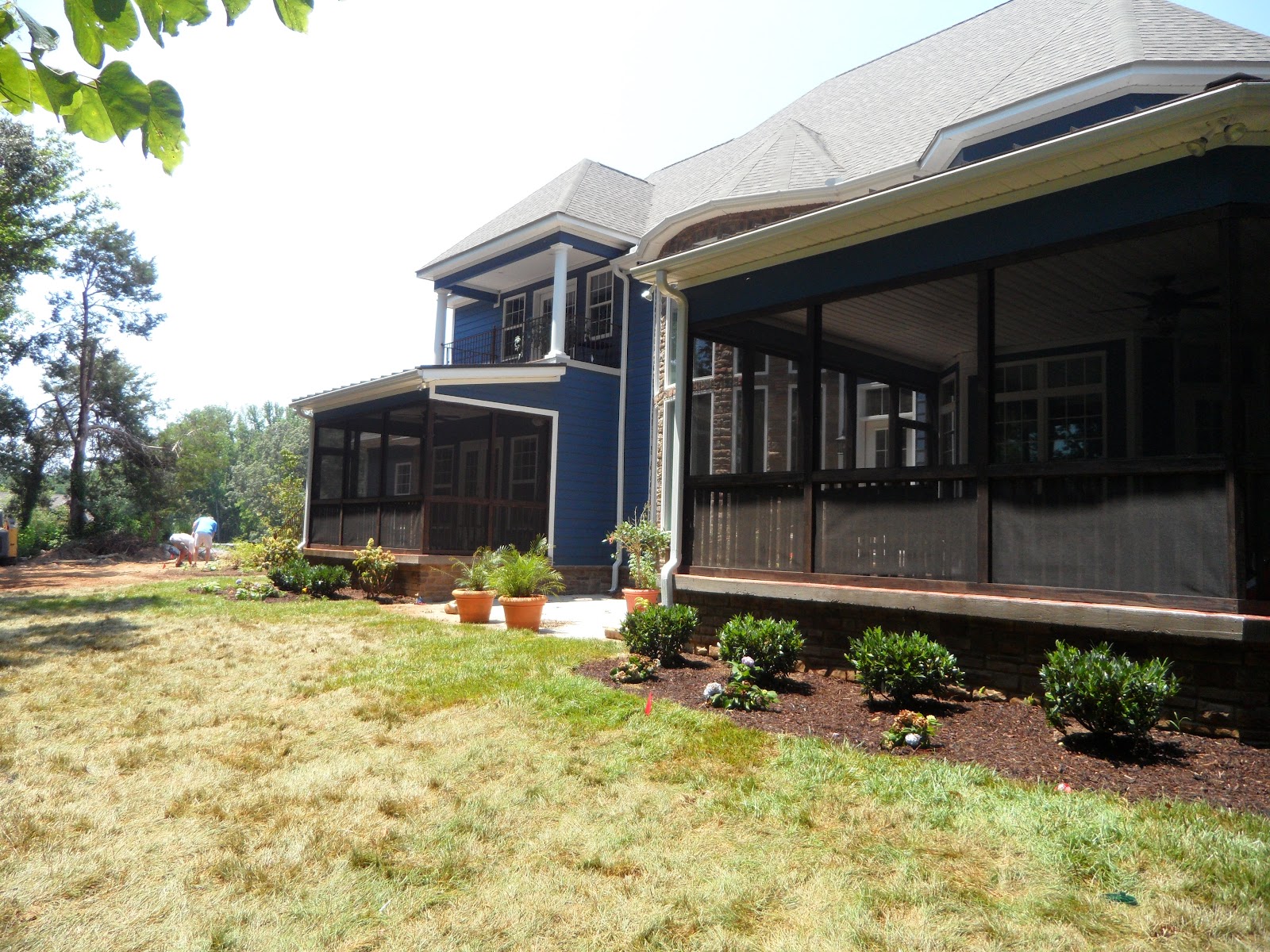 Rear exterior of a blue house with multiple screened-in porches, stone foundation accents, and a lawn with small shrubs and potted plants.