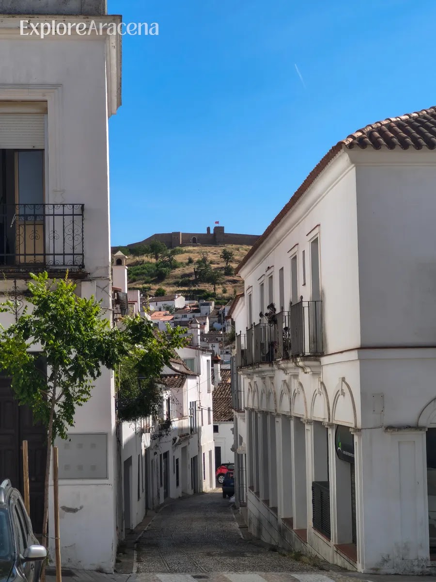 Narrow street in Aracena lined with white buildings, with the castle visible on the hill in the distance.