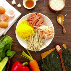 Sơ chế: Gạo dẻo nấu chín, sau đó trộn với 1 muỗng canh giấm gạo, 1 muỗng cà phê muối, 1 muỗng cà phê dầu mè. Thanh cua, chả cá, xúc xích, củ cải vàng, cà rốt, dưa leo, ớt chuông đỏ cắt sợi vừa ăn rồi đem tất cả xào sơ 2 phút. Trứng đánh tan cho thêm 1 muỗng cà phê muối, sau đó đem chiên chín rồi cắt thành sợi vừa ăn. Rau cải xanh đem luộc, tiếp đến trộn với 1 muỗng cà phê dầu mè, 1 muỗng cà phê muối, trộn đều.