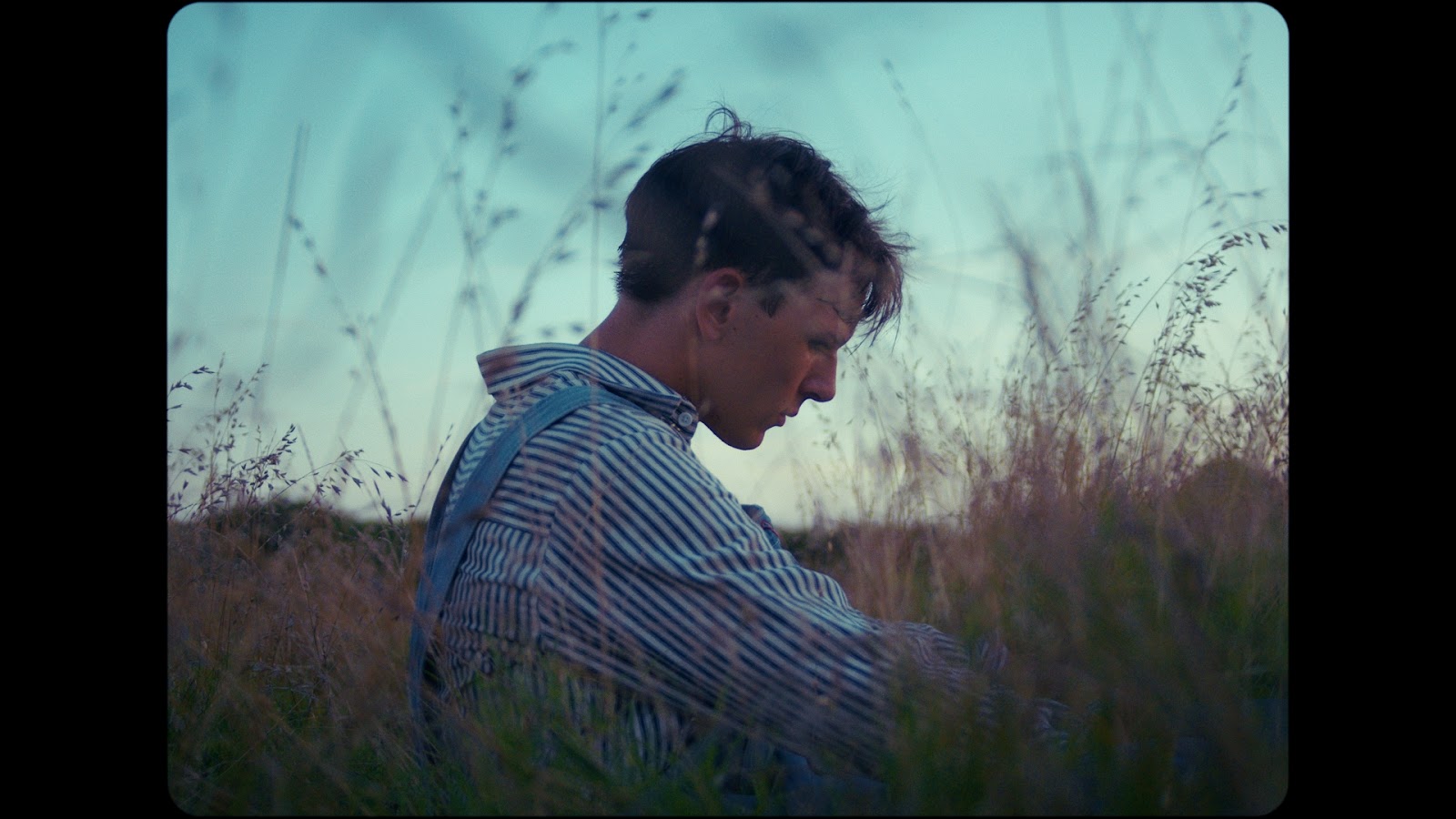 A young man in a striped work shirt sits in tall grass