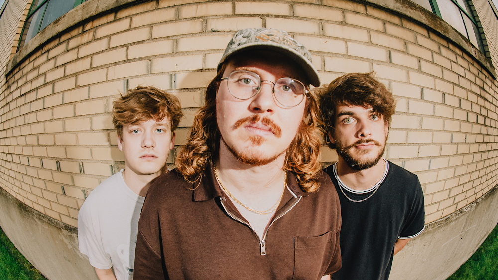 A fisheye lense photo of three men stand mostly side by side in front of a brick wall. 