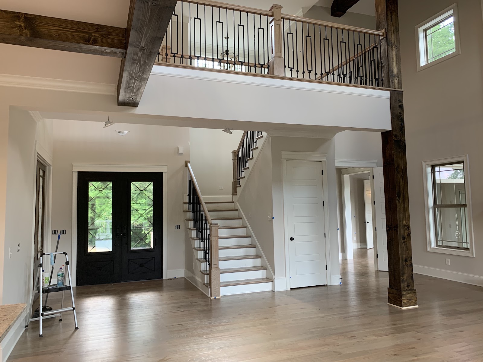 Interior foyer with light wood flooring, a staircase with dark treads, a black double front door, and a second-floor balcony with a metal railing.
